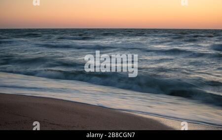 An einem warmen Sommerabend spritzen faszinierende Meereswellen entlang des Sandstrands gegen einen wunderschönen orangen Sonnenuntergang. Das Konzept der langen Sommertage durch den Stockfoto