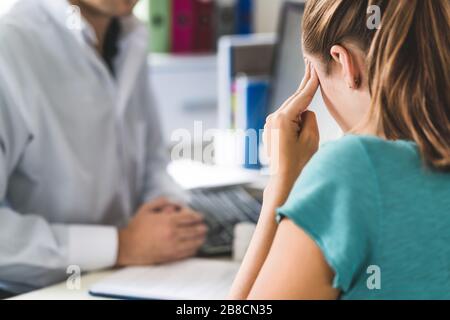 Frau, die an schlechten Kopfschmerzen oder Migräne leidet. Termin mit Arzt im Büro. Kranke und unwohl Frau mit Stress, Trauma oder Burnout. Stockfoto
