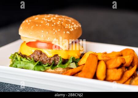 Burger Mahlzeit auf dem Teller. Köstlicher Hamburger mit saftigem Rindfleisch, schmelzender Cheddar-Käse serviert mit knusprigen Süßkartoffelfries. Hausmannskost. Stockfoto