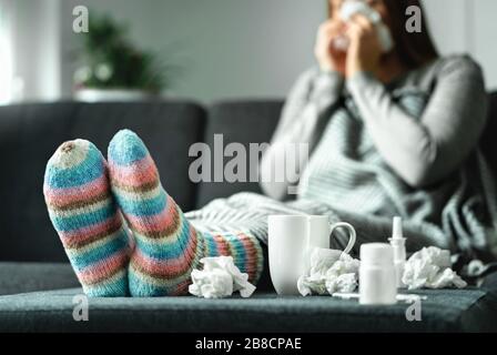 Kranke Frau mit Grippe, Kälte, Fieber und Husten, die zu Hause auf der Couch sitzt. Kranke blästen Nase und niesen mit Gewebe und Taschentuch. Wollsocken. Stockfoto