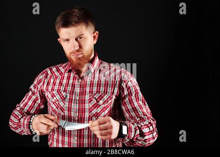 Bärtiger Rotkopfmann in einem karierten Hemd, das die Schärfe seines Messers auf schwarzem Hintergrund getestet hat. Koch bereitet sich auf die Arbeit vor. Stockfoto