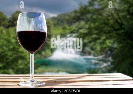 Glas Wein auf Holztisch mit großem Wasserfall verschwimmen in Kroatien. Sonniger Blick auf ein Glas Rotwein mit Blick auf den Park, Kroatien Stockfoto