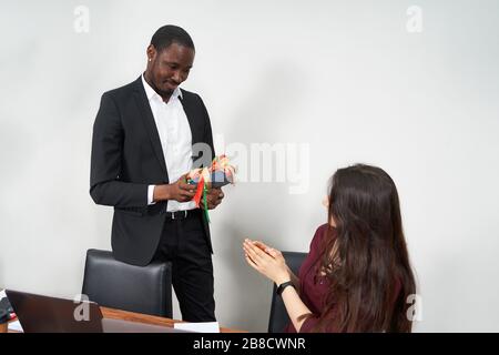 Frau, die während ihrer Arbeit im Büro ein schönes Geschenk von ihrem männlichen Kollegen erhält, Beziehungskonzept Stockfoto