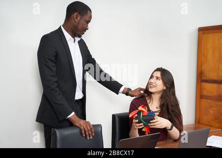 Frau, die während ihrer Arbeit im Büro ein schönes Geschenk von ihrem männlichen Kollegen erhält, Beziehungskonzept Stockfoto