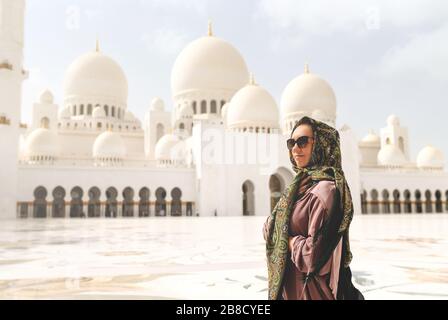 Glückliche Frau in der Scheich Zayed Grand Moschee. Touristin mit Kopftuch und Kleid in Adu Dhabi. Dame, die in einem traditionellen muslimischen Gebäude spazieren geht. Stockfoto