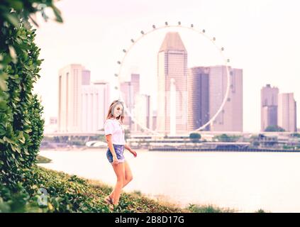 Positive trendige Frau in Jeans-Shorts, die in einem Park im Freien spazieren gehen und die Skyline der Stadt im Hintergrund sehen. Fröhlicher modischer Lebensstil. Stockfoto