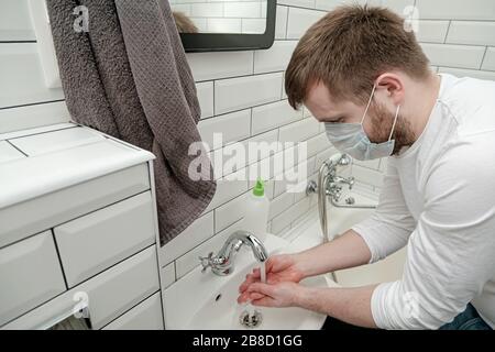 Der Mann in einer medizinischen Maske wäscht die Hände vorsichtig mit flüssiger Seife unter einem Wasserstrom, um sich vor einem gefährlichen Virus zu schützen. Stockfoto