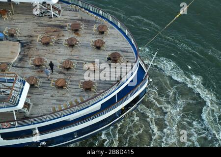 Hubschrauberblick auf das Heck des Kreuzfahrtschiffs mit Holzdeck, das von der Anlegestelle weggeschleppt wurde, mit starkem Strom im Wasser von Motoren voller Kraft Stockfoto