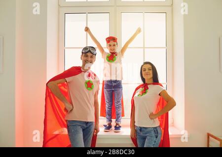 Fröhliche fröhliche Familie in Kostümen von Superhelden lachen, während sie vor dem Hintergrund eines Fensters in einem Raum drinnen steht. Stockfoto