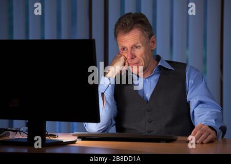 Müde gelangweilte Geschäftsmann, der nachts in einem blauen Büro einen Computermonitor betrachtet Stockfoto