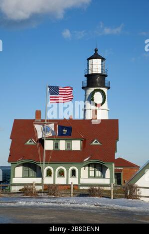 Amerikanische Flaggenwellen vor dem Leuchtturm von Portland Head, der während der Ferienzeit an einem windigen Wintertag in Maine mit einem riesigen Kranz dekoriert war. Stockfoto