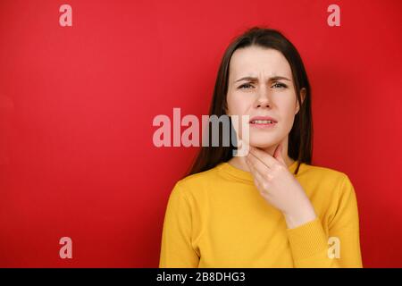 Müde junge weibliche Modell hat Halsschmerzen, ist krank und unwohl, Stirnrunzeln, spürt Schmerzen nach dem Fangen von Kälte, trägt gelben Pullover, steht auf rotem Bolzen Stockfoto