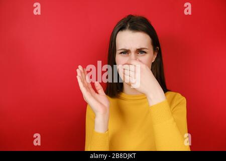 Unglückliche junge Frau bedeckt die Nase mit der Hand, riecht etwas Schreckliches, kneift die Nase, Stirnrunzeln im Unmut, gekleidet in gelben Pullover Stockfoto