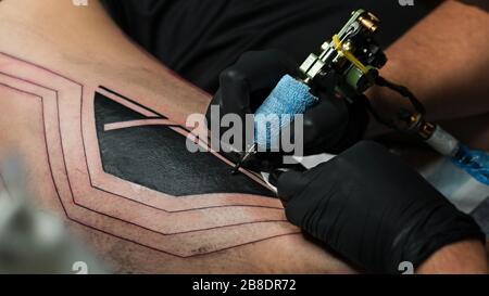 Professioneller Tattoo-Künstler macht Tattoo am Mannbein. Führt schwarze Tinte mit der Nadel von der Tattoo-Maschine in die Haut ein. Nahaufnahme der Tattoo-Maschine. Tätowierung. Erstellen eines Bildes auf dem Bein. Stockfoto