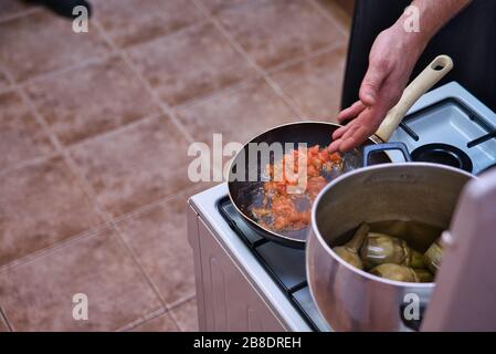 Kochen einer hausgemachten Tomatensauce in einer Bratpfanne auf einem Gasfeuer und schwelende Artischocken mit Öl und Holzkohle. Hand des erwachsenen Mannes. Stockfoto