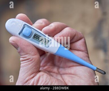 Bei der globalen Kovid-19- oder Corona-Pandemie hält die männliche Hand das klinische Thermometer mit einem Kovid-19-Text auf dem Bildschirm Stockfoto