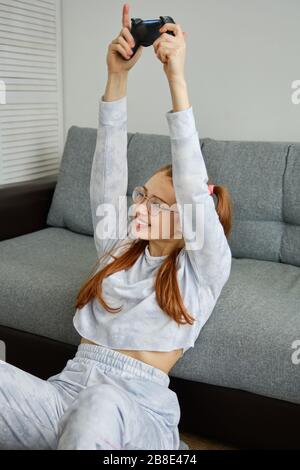 Auf dem Boden sitzt ein Rotkopfmädchen mit Brille, mit langen Schwänzen, das sich auf das Sofa lehnt und die Hände freudig von der Spielkonsole hebt Stockfoto