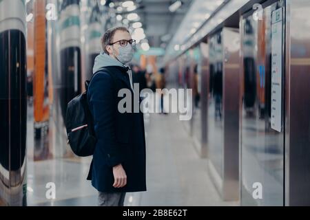 Horizontaler Schuss von Reisenden auf U-Bahn-Station, trägt medizinische Maske während Coronavirus-Zeit, wartet auf Zug, pendelt zur Arbeit mit öffentlichen Verkehrsmitteln Stockfoto