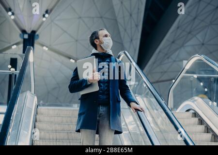 Schuss von Touristenposden auf Rolltreppe im Flughafen, kommt bei Virusausbruch aus dem Ausland nach Hause, trägt eine medizinische Schutzmaske. Evakuierter Passagier. Stockfoto