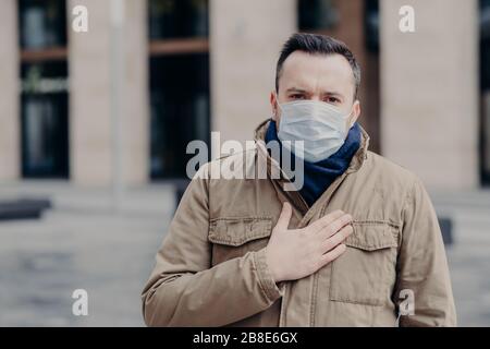 Neuartiges Coronavirus. Unzufriedener Mann hält Hand auf dem Brustkorb, hat Probleme mit der Atmung, trägt eine Schutzmaske, leidet unter einem neuen Virus, das die Lunge betrifft Stockfoto