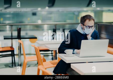 Freiberuflicher Mann trägt Gesichtsmaske, um die Ausbreitung von Coronavirus und Husten zu verhindern, verwendet Laptop für ferne Arbeit, Posen im Café mit Tasse Kaffee, Stockfoto