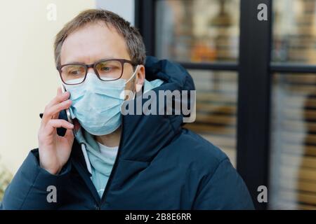 Quarantäne des Coronavirus. Trauriger verwirrter Mann in Panik wegen epidemischer Krankheit, bedeckt Gesicht mit medizinischer Maske, trägt Brille, hält Handy in der Nähe Stockfoto