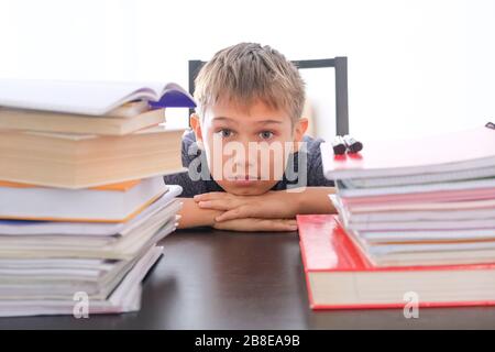 Müde enttäuschte der Schuljunge, der auf seinem Schreibtisch zu Hause zwischen Büchern, Lehrbüchern und Schulbüchern sitzt Stockfoto