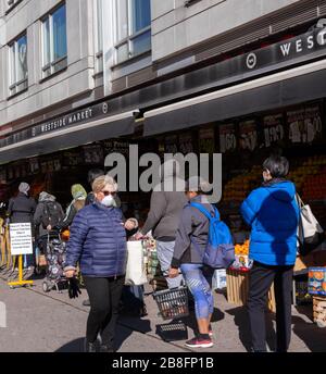 Eine kaukasische Frau, die eine Gesichtsmaske trägt, geht an Menschen vorbei, die in einer Entfernung von sechs Fuß warten, um während der Coronavirus-Pandemie in den Westside-Markt zu gelangen Stockfoto