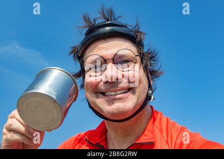 Porträt eines lächelnden Radfahrers im Retro-Helm trinkt Wasser aus Teacup. Durstiger Biker trinkt Getränk gegen blauen Himmel. Stockfoto