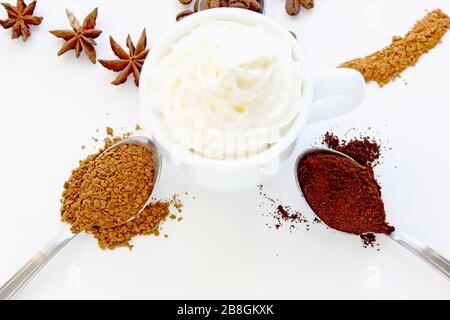 Tasse Cappuccino mit Zimt und verschütteten Kaffeebohnen. Eine Tasse Kaffee mit Schlagsahne auf dem Hintergrund verschiedener Kaffee und Gewürze. Stockfoto