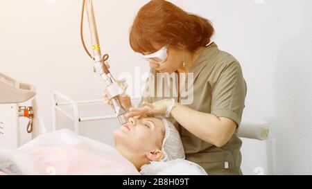 Smiling brunette Mitarbeiter in speziellen Sonnenbrillen entfernt Haare von jungen Frau Gesicht mit Laser Gerät im Beauty Salon Stockfoto