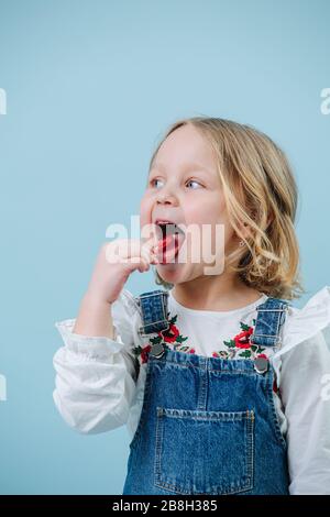 Fröhliches kleines blondes Mädchen im Jeanskleid genießt es, Lollipop-Süßigkeiten über Blau zu essen Stockfoto