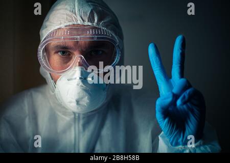 Ein Arzt mit einem müden Blick in eine Atemschutzmaske, eine Brille und einen bakteriologischen Schutzanzug zeigt ein Siegeszeichen mit einer Hand in einem blauen medizinischen Handschuh. Co Stockfoto