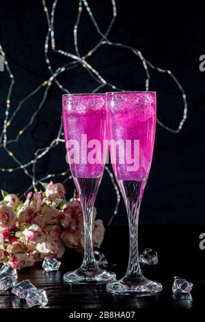 Strahlend brillantes Himbeer-Getränk. Sekt in zwei Gläsern steht auf einem Holztisch mit Eis. Für Blumen und eine Girlande. Kopierbereich Stockfoto