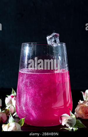 Auf einem Holztisch mit Eis steht ein funkelndes, brillantes Himbeergetränk aus Glas. Im Hintergrund stehen Rosen und eine Girlande. Kopierbereich Stockfoto