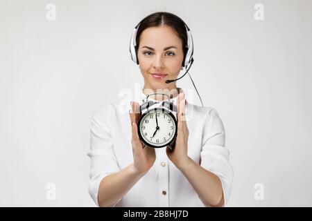 Schöne lächelnde Frau Beraterin des Callcenters in Kopfhörern, die den Wecker auf grauem Hintergrund hält. Weibliche Kundenbetreuerin mit Headset. Sparen Sie Zeit Stockfoto