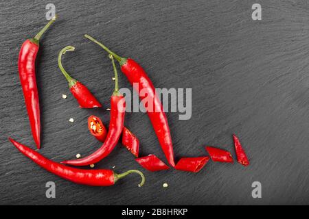 Ganze und in Scheiben geschnittene heiße rote Paprika auf einem schwarzen Schneidebrett. Ansicht von oben. Stockfoto