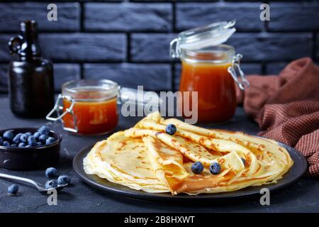 Frisch gekochte Crepes mit Blaubeeren auf einem Teller mit hausgemachtem Karamell oder Buttermilchsirup in Glasgefäßen auf einem Betontisch im Hintergrund Stockfoto