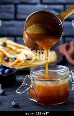 Frisch gekochter hausgemachter Buttermilchsirup gießt aus Kupfertopf zu einem Glasbecher, Nahaufnahme, vertikaler Blick Stockfoto