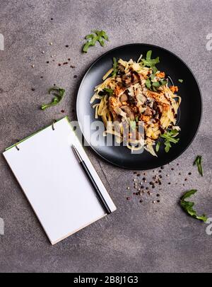 Bolognese Pasta mit Pilzen und Arugula auf schwarzem Teller gebuste Balsamico-Sauce. Ein Buch zum Schreiben von Rezepten liegt auf einem Tisch. Stockfoto