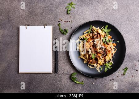 Bolognese Pasta mit Pilzen und Arugula auf schwarzem Teller gebuste Balsamico-Sauce. Ein Buch zum Schreiben von Rezepten liegt auf einem Tisch. Stockfoto