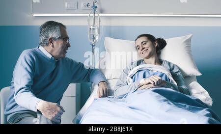 Mann, der ihre Tochter im Krankenhaus besucht, die Frau liegt im Bett und lächelt Stockfoto