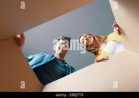 Glückliches Paar öffnet einen Pappkarton und blickt nach innen, sie lächeln und sind fröhlich Stockfoto
