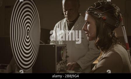 Wissenschaftler, die Hypnose-Experimente im Labor im Vintage-Stil durchführen, starrt eine Frau auf die rotierende Spirale und wird hypnotisiert Stockfoto