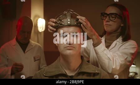 Wissenschaftler, die in einem Labor im Vintage-Stil an einem menschlichen Gehirn experimentieren, trägt ein Mann einen Sci-Fi-Helm und Forscher testen ihn Stockfoto