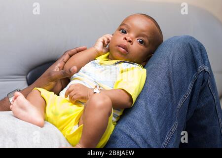 Neugeborenes Baby, das auf den Beinen seines Vaters auf der Couch zu Hause liegt und dabei die Kamera beobachtet. Stockfoto