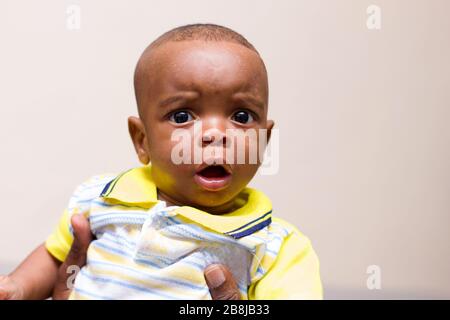 Nahaufnahme des Babys in den Armen seines Vaters, der zu Hause auf der Couch sitzt und dabei die Kamera überrascht beobachtet Stockfoto