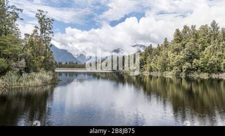 Landschaft mit Bergen, Wolken und grünem Wald, der in Gewässern reflektiert wird, die in hellem bewölktem Licht am Matheson Lake, Westküste, Südinsel, Neuseeland geschossen werden Stockfoto