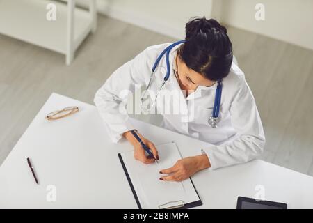 Selbstbewusste Frau Ärztin Kinderärztin schreibt in einer Zwischenablage, die an einem Tisch in einem weißen Büro des Krankenhauses sitzt Stockfoto