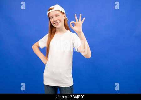 Charmantes rothaariges Teenager-Mädchen in einem weißen T-Shirt zeigt einen leeren Druckraum auf blauem Studiohintergrund Stockfoto